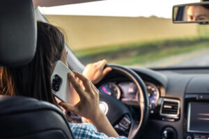 Vista desde el asiento trasero de un coche de una conductora con una camisa de cuadros, que sujeta un teléfono inteligente blanco contra su oreja izquierda mientras conduce en una carretera recta. ¿Cómo se puede probar la conducción distraída después de un accidente automovilístico?