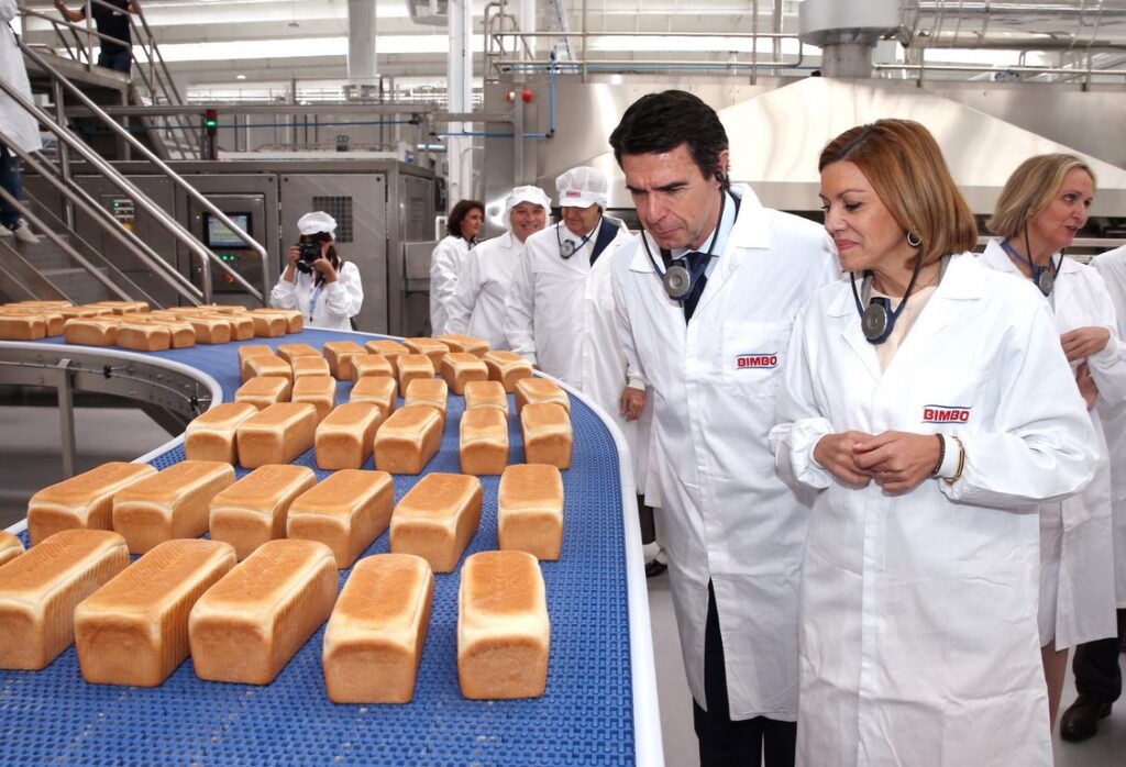 A woman and a man, wearing white lab coats with the Bimbo logo, observe baked loaves of bread on a blue conveyor belt inside a factory.