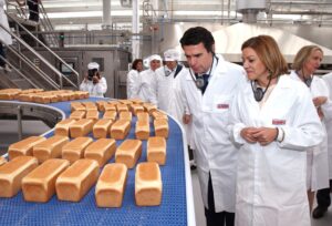 A woman and a man, wearing white lab coats with the Bimbo logo, observe baked loaves of bread on a blue conveyor belt inside a factory.