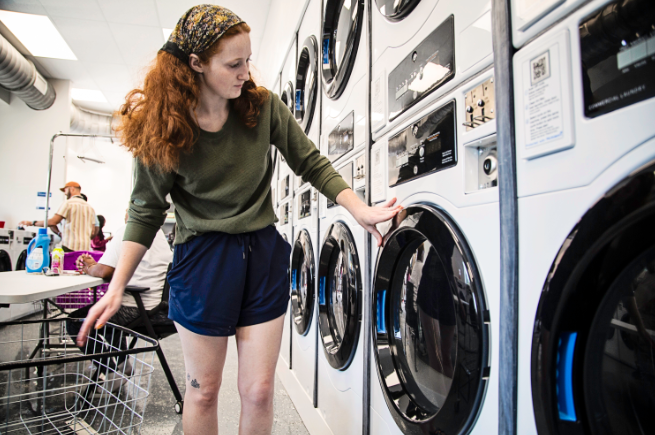 A woman with red hair, wearing a bandana and blue shorts, checks the control panel of a white front-loading washing machine in an industrial laundromat.
