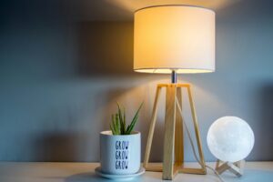 Una lámpara de mesa moderna con base de madera estilo trípode y pantalla blanca cilíndrica encendida. A un lado, una maceta con una planta pequeña y al otro, una lámpara de luna.