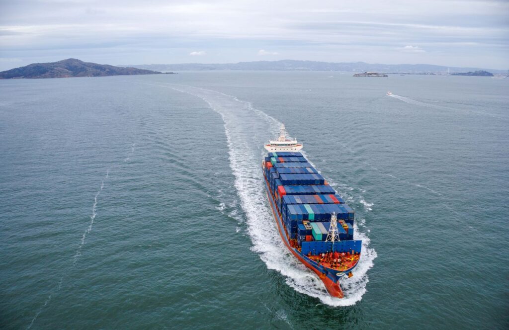 Aerial view of a large container ship loaded with blue and red containers, sailing on the open sea, leaving a white spray of foam, with hills on the horizon. Photo: Robert So, via Pexels. Maritime infrastructure and efficiency at North American ports represent the new strategic frontier for ensuring operational resilience and logistical competitiveness within the framework of the USMCA.