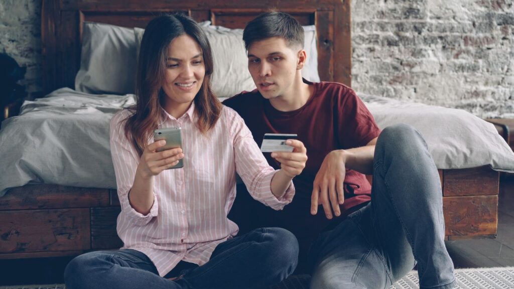 Una pareja sentada en el suelo de un dormitorio; la mujer sostiene un celular y el hombre una tarjeta de crédito, ambos mirando el teléfono para realizar un pago online.