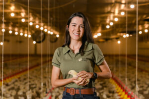 A woman smiles in a modern poultry barn, holding a chick. Her T-shirt reads “Vida Granja,” illustrating the high-tech production of Brazilian chicken for export.