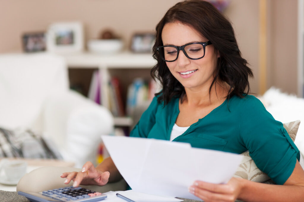 Una mujer con lentes sonríe mientras revisa documentos en papel y utiliza una calculadora, sentada cómodamente en un sofá de su hogar con estanterías de fondo. Evita autodeterminar tus impuestos: el peligro de automedicarse fiscalmente.