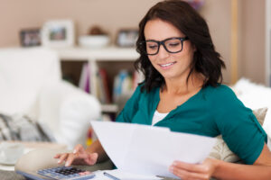 Una mujer con lentes sonríe mientras revisa documentos en papel y utiliza una calculadora, sentada cómodamente en un sofá de su hogar con estanterías de fondo. Evita autodeterminar tus impuestos: el peligro de automedicarse fiscalmente.