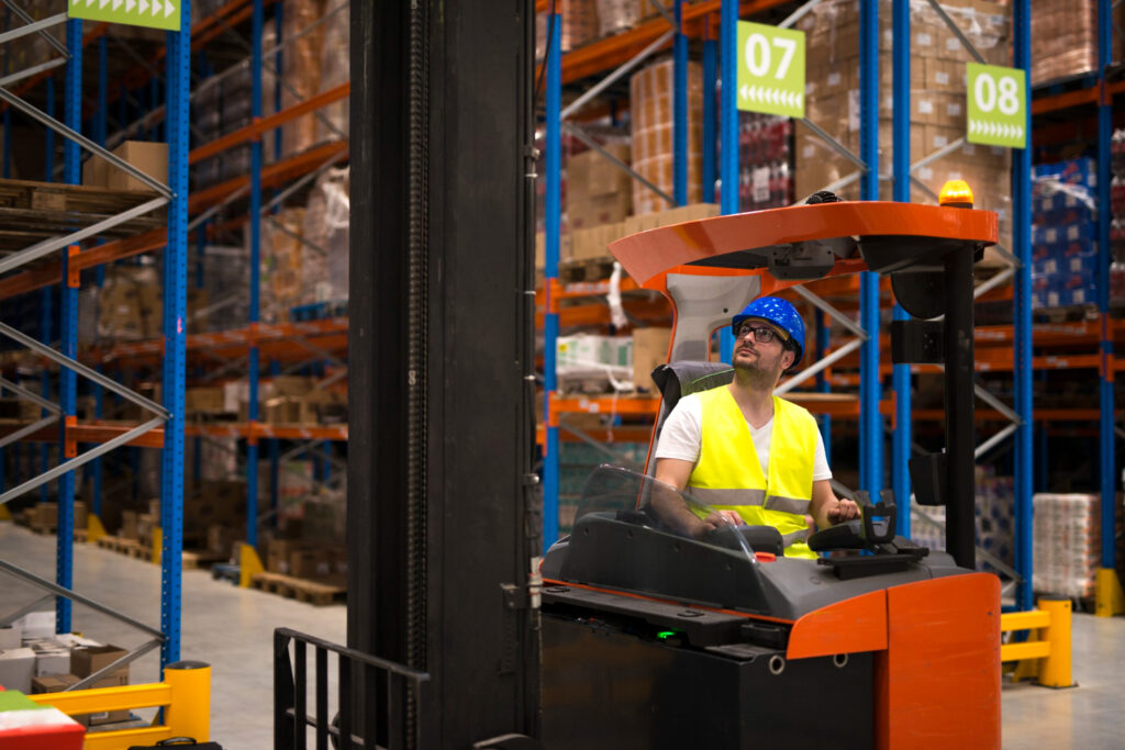 Operador de montacargas con equipo de seguridad mira hacia arriba dentro de un pasillo estrecho con estanterías industriales altas repletas de cajas de inventario organizado. Estrategias para maximizar el uso del espacio vertical en almacenes pequeños.