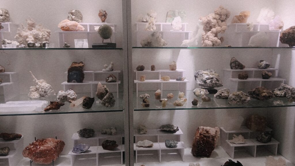 Close-up of a multi-tiered glass display case showcasing dozens of mineral specimens in white, gray, brown, and purple on transparent acrylic stands, illuminated by bright lighting.