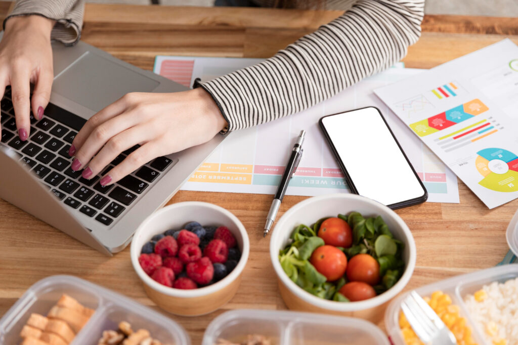 Vista superior de una persona escribiendo en una laptop sobre un escritorio de madera, rodeada por un teléfono inteligente, reportes impresos y recipientes con alimentos frescos y saludables.