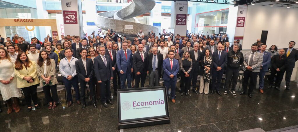 A large crowd poses for a group photo in a large, modern atrium. In the center is a small screen displaying the logo of the Ministry of Economy.