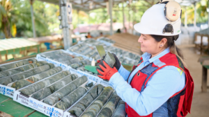 A geologist wearing a hard hat, safety vest, and gloves examines a rock core extracted from a core box at a mining facility for critical mineral analysis.