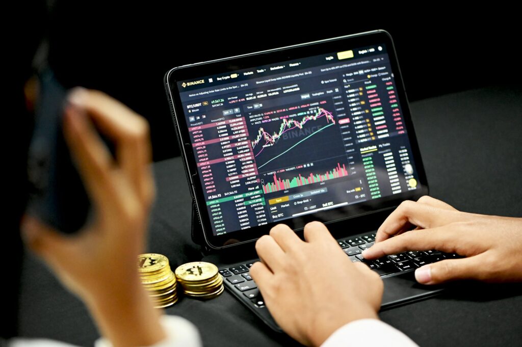 Inversor analizando la interfaz financiera de Binance en una tableta con gráficos de velas y profundidad de mercado, mientras utiliza un teléfono y tiene monedas físicas de Bitcoin cerca.