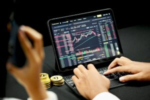 Inversor analizando la interfaz financiera de Binance en una tableta con gráficos de velas y profundidad de mercado, mientras utiliza un teléfono y tiene monedas físicas de Bitcoin cerca.