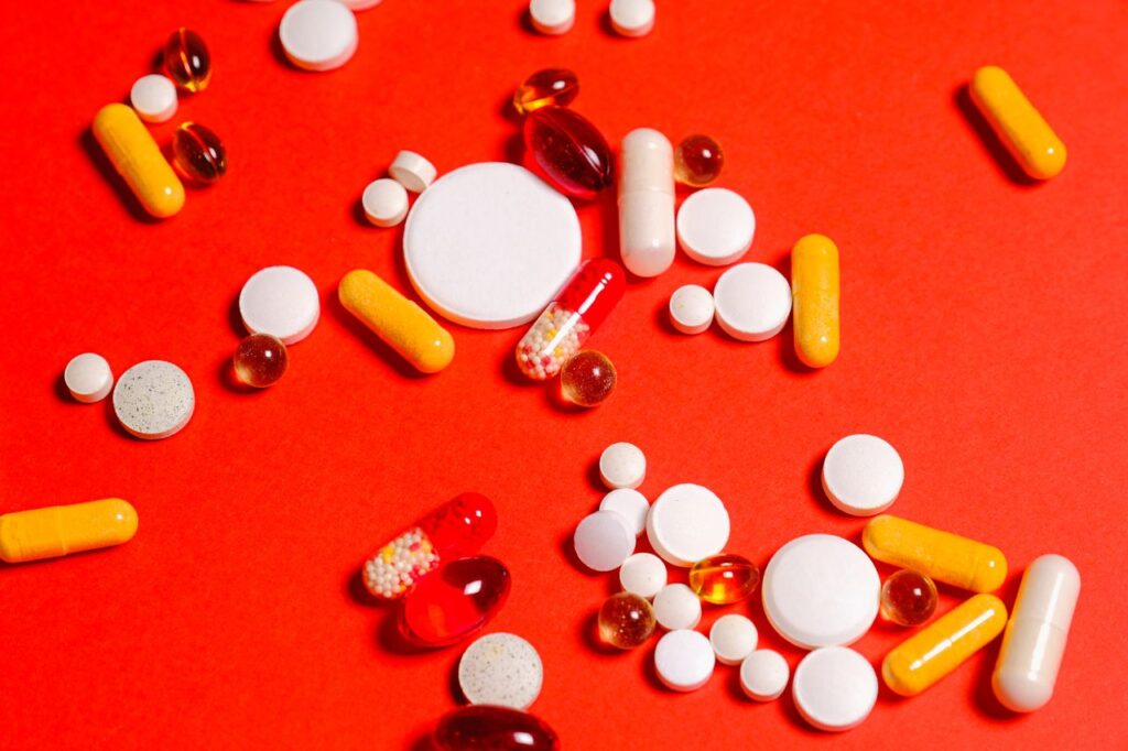 A variety of pills, white tablets, and yellow and red pharmaceutical capsules scattered in clusters on a bright red background, in a detailed overhead view.