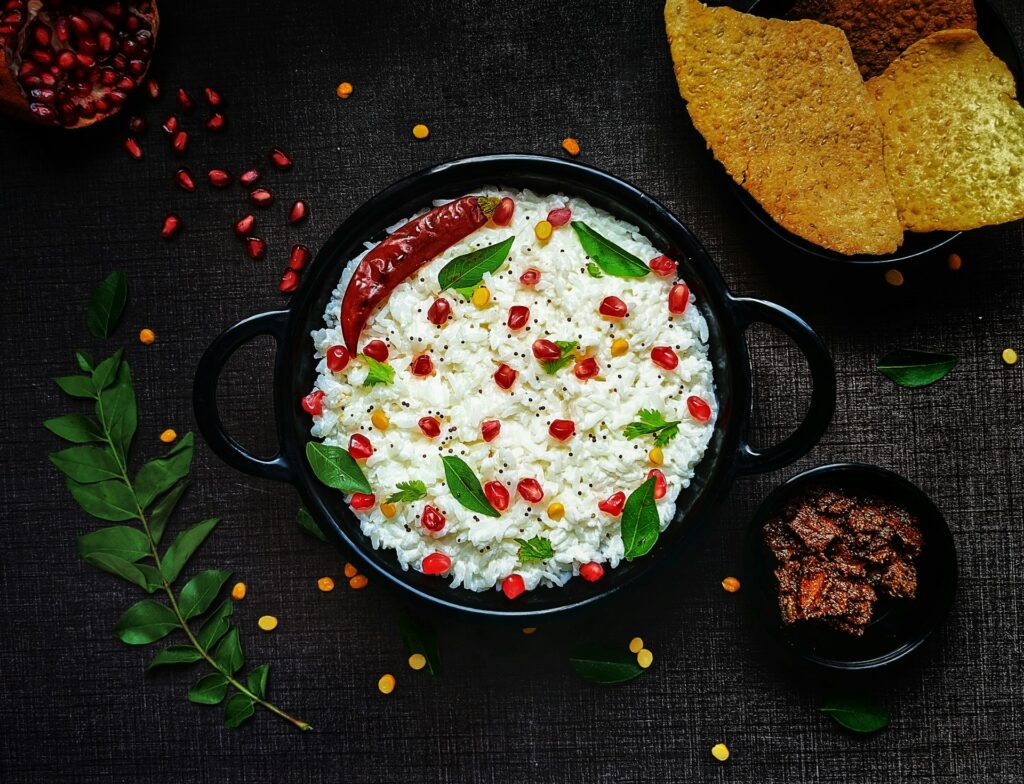Vista superior de uma tigela preta de arroz com romã e pimenta, rodeada por papadums, picles de manga e folhas de curry sobre fundo preto.