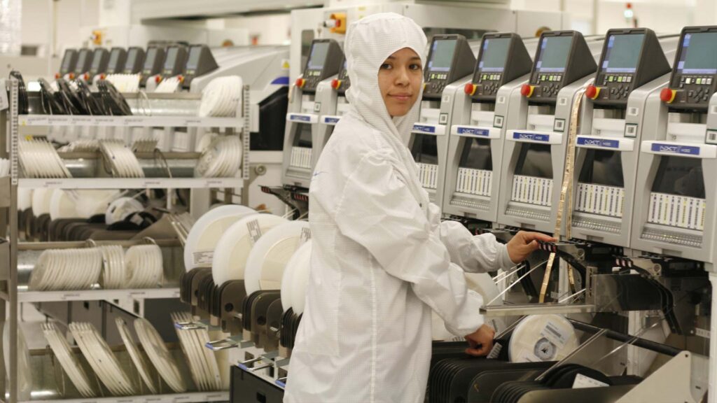 Operaria vestida con traje de protección esterilizado opera maquinaria industrial especializada de montaje electrónico en una planta de manufactura avanzada con componentes de precisión e instrumentación automatizada.