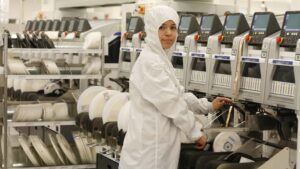 Operaria vestida con traje de protección esterilizado opera maquinaria industrial especializada de montaje electrónico en una planta de manufactura avanzada con componentes de precisión e instrumentación automatizada.
