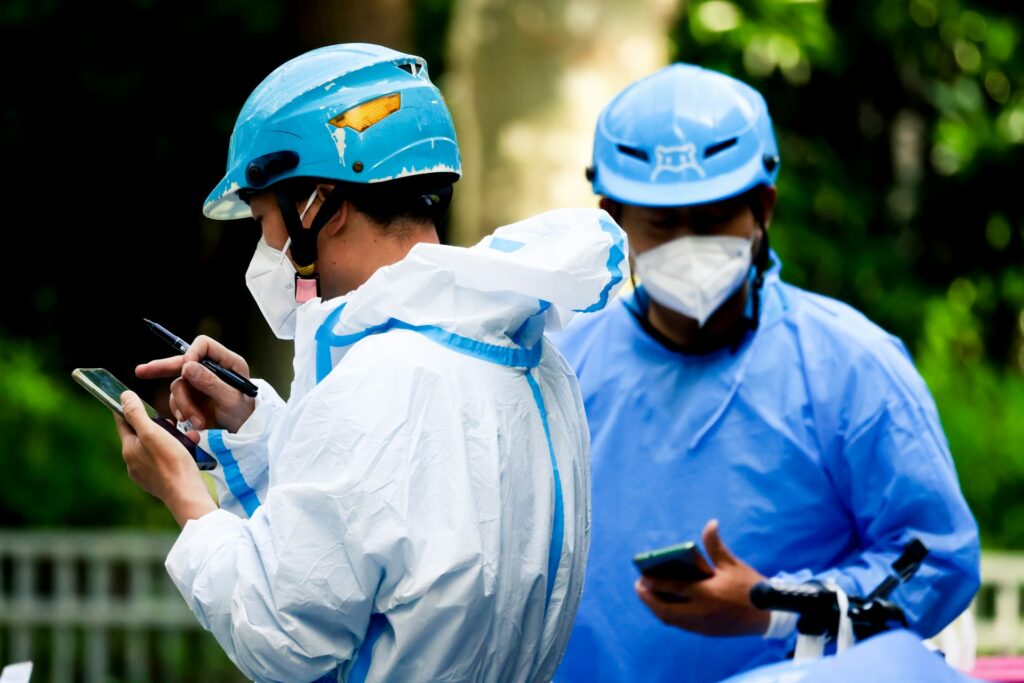 Dos personas con casco y mascarilla usan dispositivos móviles, vestidas con equipo de protección, en un entorno exterior desenfocado, ilustrando prevención sanitaria y seguridad durante viajes internacionales.