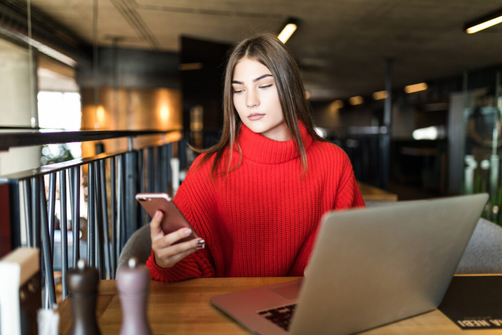 Una mujer joven con suéter rojo observa fijamente la pantalla de su teléfono móvil mientras trabaja en una mesa con una computadora portátil abierta en una cafetería.