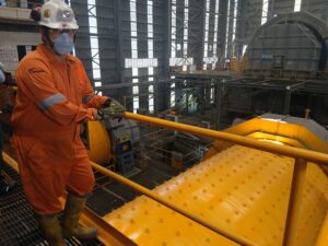 A worker wearing a safety helmet, mask, and orange coveralls supervises heavy machinery and industrial mills at a mining processing plant in Mexico.