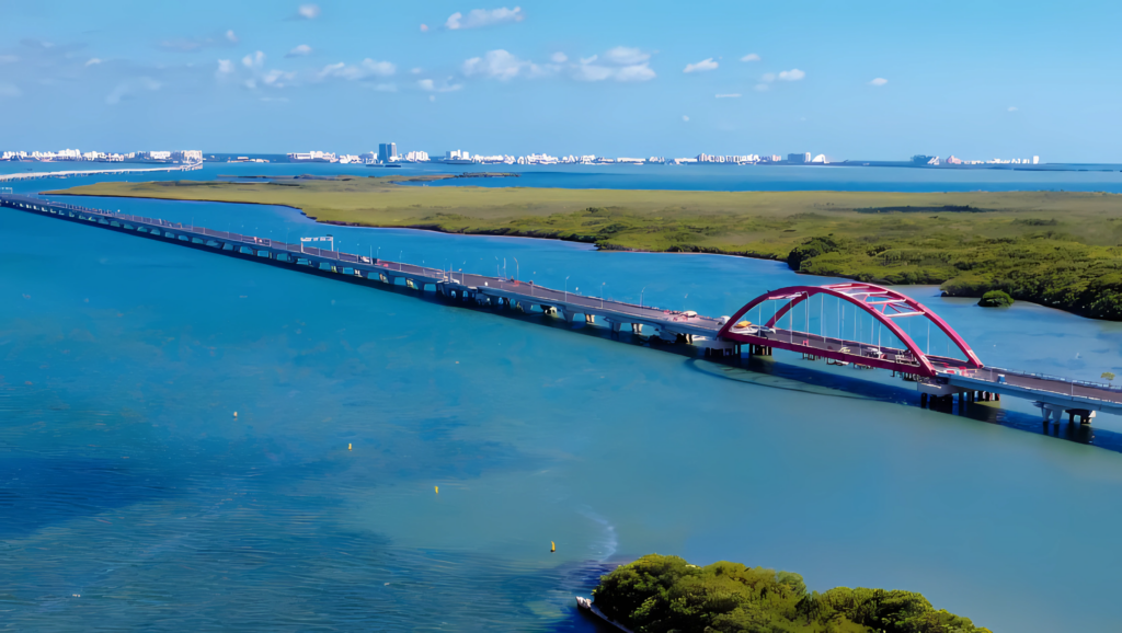 Vista aérea panorâmica da Ponte Rodoviária Nichupté a atravessar a lagoa turquesa, destacando a sua estrutura elevada e um característico arco metálico vermelho rodeado por vegetação de mangue.
