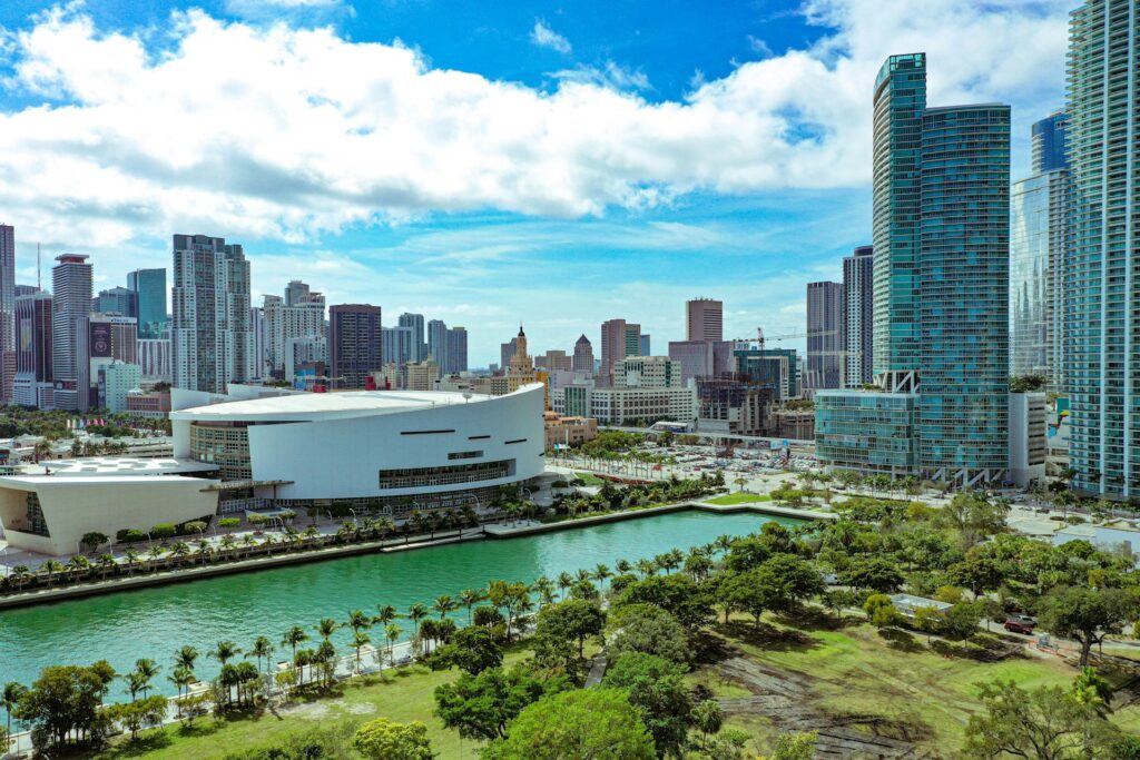 Vista aérea del centro urbano de Miami con rascacielos modernos, el estadio Kaseya Center frente a la bahía de Biscayne, áreas verdes y un canal con agua turquesa bajo cielo soleado. ¿Por qué los inversionistas mexicanos están blindando su patrimonio con bienes raíces en Miami?
