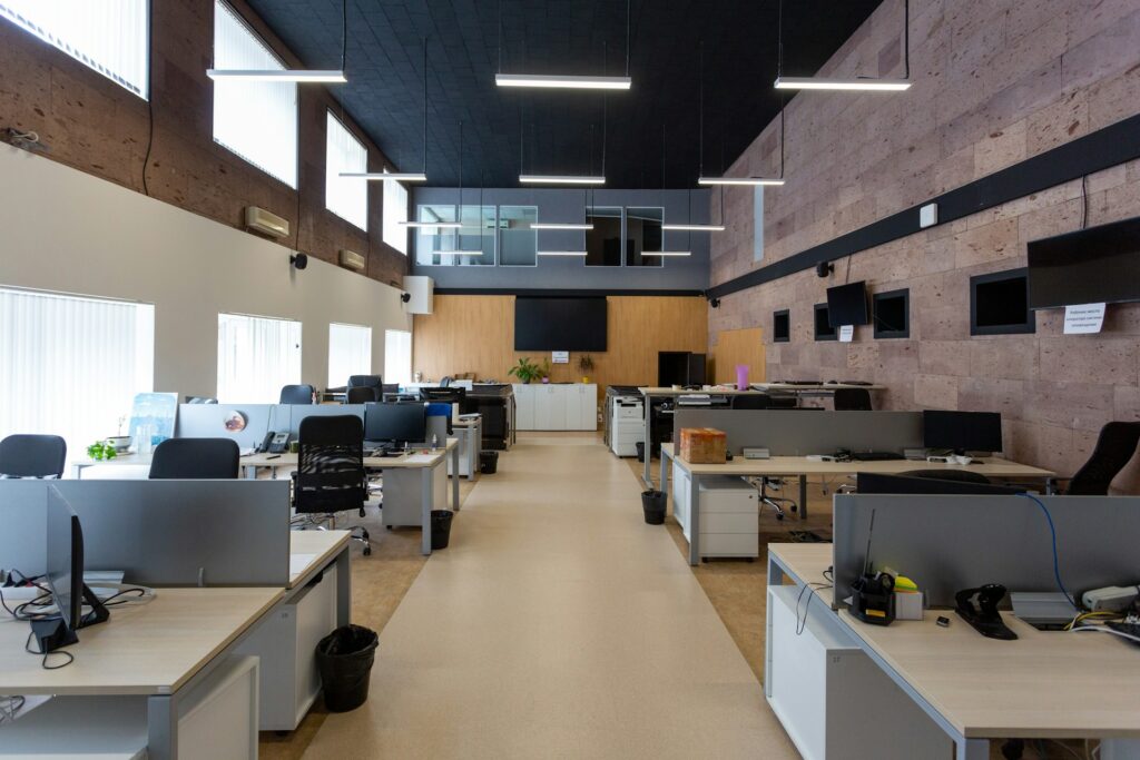 Amplio espacio de trabajo corporativo con múltiples escritorios y computadoras, iluminación LED suspendida, techo oscuro, mamparas divisorias y muros revestidos con acabado de piedra natural.