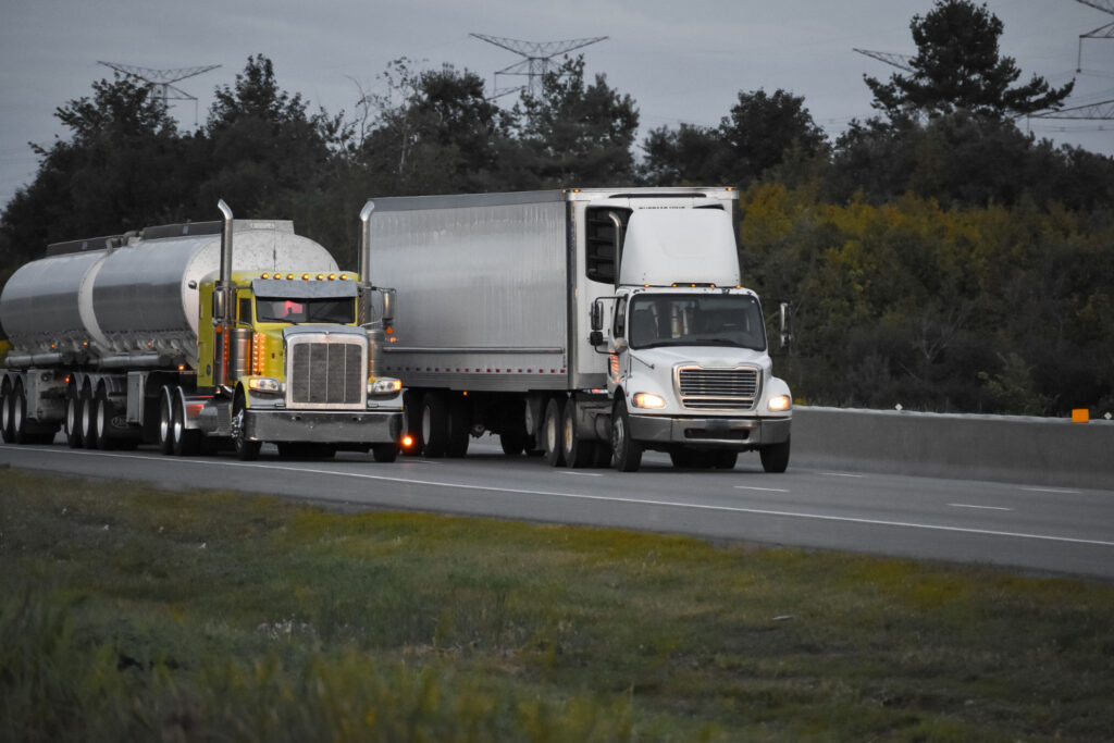 Two cargo trucks—a yellow tanker and a white trailer—travel along a highway, symbolizing logistics, heavy haulage, and trade integration in North America.