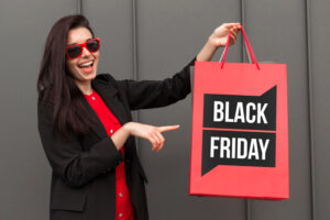 Una joven sonriente con lentes de sol rojos y chaqueta negra sostiene una bolsa de compras roja que dice «BLACK FRIDAY» en texto negro y blanco sobre un fondo gris.