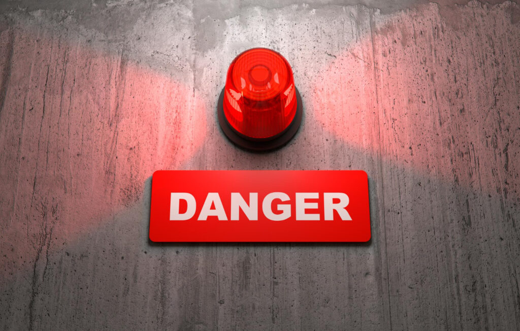 Una luz de alarma roja encendida emite un destello brillante sobre una superficie de cemento texturizado. Debajo de la luz, hay un cartel rectangular rojo con la palabra "DANGER" (PELIGRO) escrita en letras blancas mayúsculas. Infraestructura empresarial y prevención: inversiones clave para reducir riesgos.