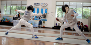 Dos tiradores de espada en plena acción de combate sobre una pista de esgrima metálica, con la tiradora de la izquierda realizando un a fondo mientras el de la derecha mantiene la distancia. El doble toque como táctica consciente en la espada.